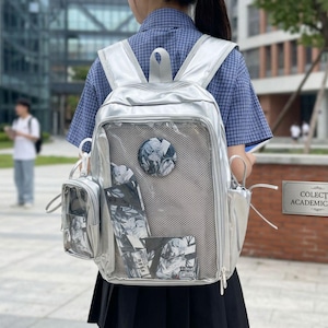 Könnte beinhalten: Ein metallisch silberner Rucksack mit einer großen Fronttasche aus Netzstoff, einem runden Anstecker mit einer Anime-Illustration und einer abnehmbaren kleinen durchsichtigen Tasche an der Seite sowie mehreren Reißverschlussfächern.