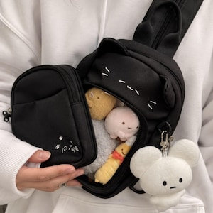 Puede incluir: Una mochila bandolera negra con orejas de gato en la parte superior, rasgos faciales de gato bordados y un bolsillo frontal pequeño con huellas. El compartimento principal está abierto, mostrando varios peluches y un colgante de felpa blanco.