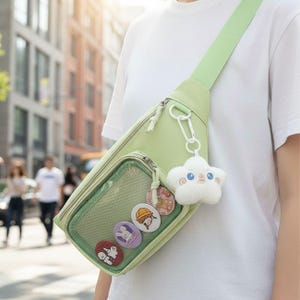 Peut inclure: Un sac bandoulière vert clair avec une poche transparente affichant des épingles rondes colorées. Le sac a une sangle assortie et un porte-clés blanc en forme de nuage. Le sac est porté en travers du corps.