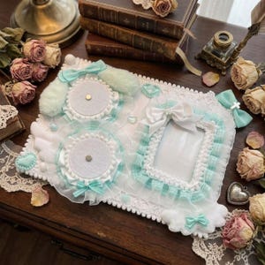 Puede incluir: Un tablero de exhibición rectangular blanco que presenta dos almohadillas circulares cubiertas de fieltro con alfileres metálicos centrales, un marco rectangular central y volantes, lazos y pequeños adornos en forma de corazón de color verde menta y blanco.