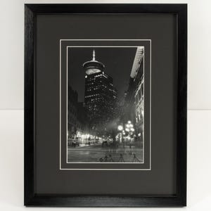 Pode incluir: Fotografia emoldurada a preto e branco de uma rua da cidade à noite. A imagem apresenta um edifício alto com uma torre distinta, candeeiros de rua e figuras desfocadas. A moldura é preta com uma passe-partout branca.