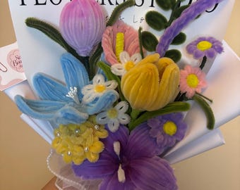 May include: A vibrant bouquet of handcrafted flowers in various colours. The arrangement includes blue, purple, yellow, pink, and white flowers, with green stems and leaves. The bouquet is wrapped in white paper with the text "FLOWERS STUDIO".