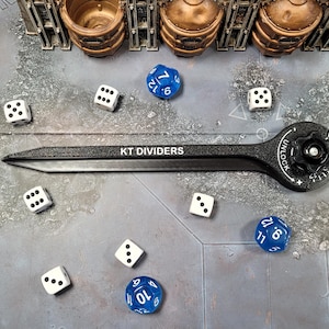 May include: A black KT Dividers tool with a dial and the word "UNLOCK" is surrounded by white dice and blue dice. The dice are scattered on a grey surface with miniature terrain pieces.