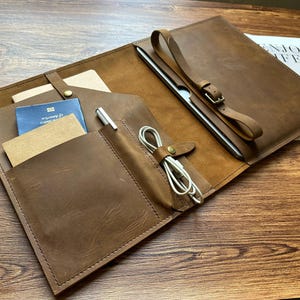 May include: Open brown leather folio with multiple pockets, holding a passport, notepad, pen, and charging cable. A laptop is secured with a strap. The folio is on a wooden surface.