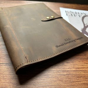 Puede incluir: Un diario de cuero marrón con un cierre de solapa asegurado por dos tachuelas de color latón. El diario está grabado con las iniciales "C.L.L." y la frase "Should Something Happen". Una revista con las palabras "Enjoy Life" está en el fondo.