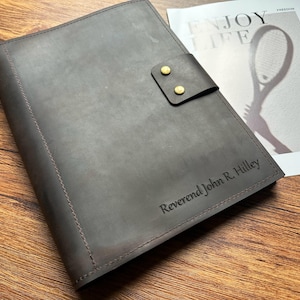 Może przedstawiać: Szary skórzany notatnik z klapką zabezpieczoną dwoma ćwiekami w kolorze mosiądzu. Notatnik ma szyte krawędzie, a na okładce wytłoczono imię i nazwisko "Reverend John R. Hilley". W tle magazyn ze słowami "ENJOY LIFE".