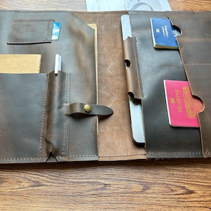 May include: Open brown leather folio with multiple pockets. Features a pen holder, card slot, and slots for a passport and a small notebook. The folio is open, revealing the interior compartments and a pen.