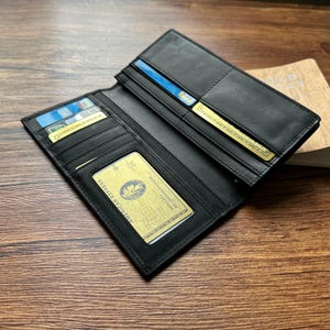 Könnte beinhalten: Offene schwarze Ledergeldbörse mit mehreren Kartenfächern, darunter ein transparentes Ausweisfach. Die Geldbörse enthält Kreditkarten und eine American Express Karte. Die Geldbörse ist geöffnet und zeigt ihr Inneres, sie liegt auf einer Holzoberfläche.