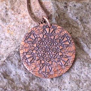 May include: Copper-colored pendant necklace with a detailed geometric pattern. The circular pendant hangs from a matching chain. The intricate design features interconnected shapes and lines, creating a textured surface. The necklace is displayed on a textured, neutral-toned surface.