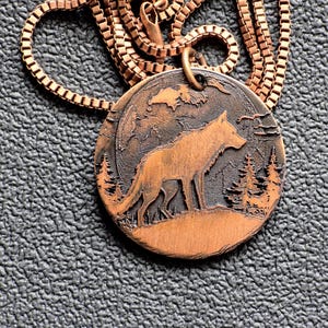 May include: Copper-colored pendant necklace featuring a detailed wolf silhouette standing on a hill, set against a backdrop of trees and a night sky. The pendant is round and hangs from a matching chain.