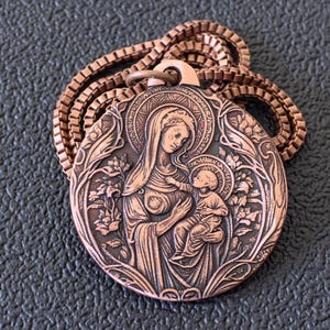 Peut inclure: Un collier pendentif de couleur cuivrée avec un relief détaillé de la Vierge Marie et de l'enfant, entouré d'éléments floraux et foliacés. Le pendentif est rond et suspendu à une chaîne de type boîte assortie.