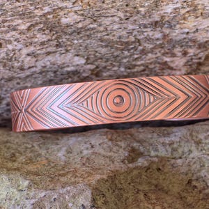 Könnte beinhalten: Ein kupferfarbenes Armband mit einem eingravierten geometrischen Design. Das Armband hat ein zentrales kreisförmiges Muster, das von winkligen Linien und Formen umgeben ist. Das Armband ist breit und hat eine polierte Oberfläche.