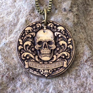 May include: A bronze-colored pendant necklace featuring a detailed skull design. The round pendant has the words "MEMENTO MORI" on a banner. The skull is surrounded by ornate scrollwork and smaller skulls.