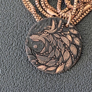 May include: A copper-colored pendant necklace featuring a detailed wolf design. The pendant is round with an intricate pattern of leaves and dots, and is suspended from a matching chain. The wolf's face is visible in the design.