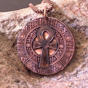 Op de afbeelding: Een cirkelvormige koperkleurige hanger met een centraal ankh-symbool, omringd door een gedetailleerde rand van diverse geëtste hiëroglief-achtige iconen. De hanger hangt aan een bijpassende koperkleurige box-schakelketting.