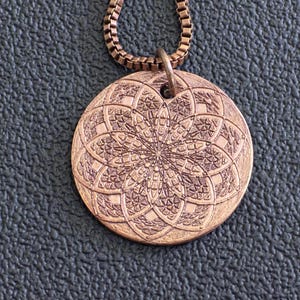 Peut inclure: Un collier pendentif de couleur cuivre avec un motif floral détaillé. Le pendentif rond est suspendu à une chaîne assortie. Le motif complexe est en relief sur la surface du pendentif, créant un effet texturé. Le collier est présenté sur une surface texturée gris foncé.