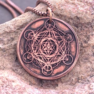 May include: A circular copper-toned pendant featuring an intricate engraved geometric pattern known as Metatron's Cube, suspended from a matching box chain. The metal surface displays a weathered, dark patina within the recessed lines.