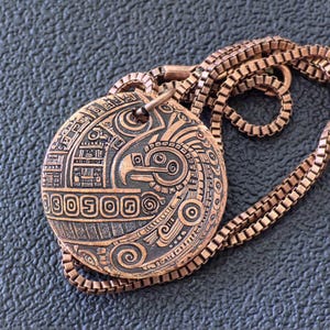 May include: A copper-colored pendant necklace with an intricate engraved design featuring an eagle or bird motif. The pendant is oval-shaped and hangs from a matching square-link chain. The design includes geometric patterns and symbols.