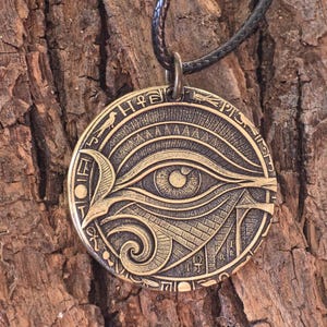 May include: A bronze-colored pendant featuring the Eye of Horus design, with intricate details and hieroglyphic symbols around the edge. The pendant hangs from a black leather cord, set against a textured wood background.