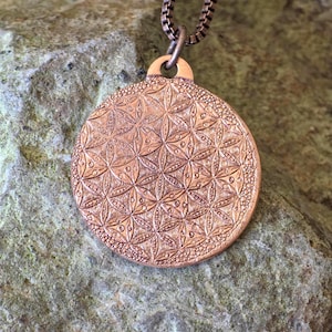 May include: A copper-colored pendant with a detailed floral pattern. The round pendant is suspended from a chain. The pendant's surface features an intricate design of repeating flower-like shapes and small dots, creating a textured appearance.