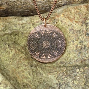 Puede incluir: Collar con colgante de color cobre con un diseño geométrico detallado. El colgante circular presenta un intrincado patrón de líneas y formas, suspendido de una cadena a juego. El colgante mide aproximadamente 3,8 cm de diámetro.