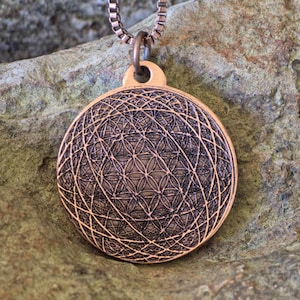Pode incluir: Um colar com pingente de cor cobre com um design geométrico. O pingente redondo apresenta um padrão detalhado de linhas e formas que se cruzam, suspenso por uma corrente correspondente. O pingente tem aproximadamente 3,8 cm de diâmetro.