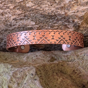 May include: A copper cuff bracelet with an etched geometric pattern. The bracelet has a rectangular shape with a series of triangles and zigzags. The bracelet is displayed on a rock surface.