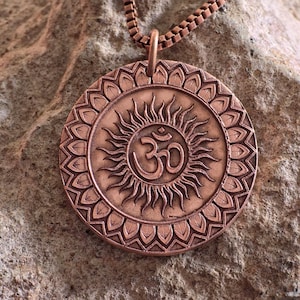 May include: Copper-colored pendant necklace featuring a circular design with a sunburst pattern and the Om symbol at the center. The pendant is suspended from a matching copper chain, set against a textured stone background.