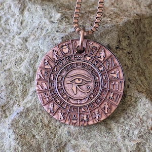 May include: A copper-coloured pendant with an Egyptian Eye of Horus design. The round pendant features hieroglyphs and symbols around the eye. The pendant hangs from a matching copper-coloured chain. The pendant is approximately 3.8 cm in diameter.