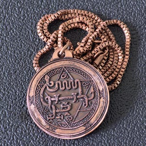 May include: A copper-colored pendant necklace with a detailed circular charm. The charm features intricate symbols and text within a star and geometric design. The chain is a matching copper color, and the necklace rests on a dark surface.