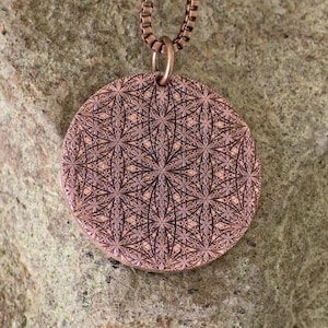 Pode incluir: Um colar com pingente de cor de cobre com um padrão floral simétrico e detalhado. O pingente redondo pende de uma corrente combinando. O design intrincado é gravado no metal, criando uma superfície texturizada. O colar é exibido em um fundo de pedra texturizada.