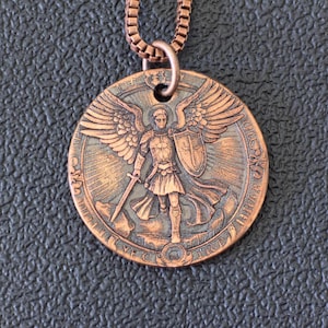 Könnte beinhalten: Kupferfarbene Halskette mit Anhänger. Der runde Anhänger zeigt ein detailliertes Relief eines Engels mit Flügeln, der ein Schwert und einen Schild hält. Der Anhänger hat einen verzierten Rand und hängt an einer passenden Kette.