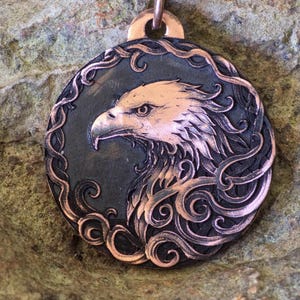 May include: A copper-toned pendant featuring a detailed profile of an eagle's head. The eagle is set against a dark background within a circular frame with ornate, swirling designs. The pendant hangs from a black cord.