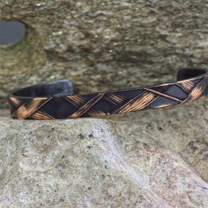 Può includere: Un bracciale a polsino in rame fatto a mano con un design intrecciato. Il bracciale presenta una finitura scura e ossidata con riflessi di rame a contrasto. Il design a polsino aperto consente di indossarlo facilmente. Il bracciale è largo circa 1,9 cm.
