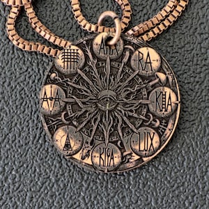 May include: A bronze pendant necklace with a detailed circular pendant. The pendant has an eye at the centre, surrounded by radiating lines and symbols. The chain is a matching bronze colour.