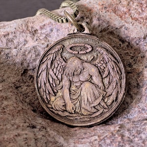 Peut inclure: Un pendentif circulaire de couleur bronze présentant un relief sculpté représentant une figure ailée agenouillée, la tête inclinée sous une auréole. Le pendentif est suspendu à une chaîne carrée assortie, avec une finition texturée et patinée.