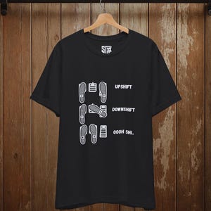 Puede incluir: Camiseta negra con un gráfico blanco de una secuencia de pie y pedal, con las palabras "UPSHIFT", "DOWNSHIFT" y "OOOH SHL." La camiseta está colgada sobre un fondo de madera.