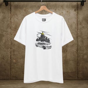 Puede incluir: Camiseta blanca con un gráfico en blanco y negro de un coche y un edificio con nubes. La camiseta tiene el texto "STX" en la parte superior. El coche está en primer plano y el edificio en segundo plano.