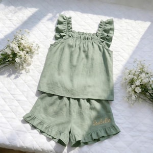 Könnte beinhalten: Salbeigrünes zweiteiliges Pyjama-Set. Das Oberteil hat Rüschenträger und einen Rüschenausschnitt, und die Shorts haben einen Rüschensaum. Der Name "Isabella" ist in Goldfaden auf die Shorts gestickt. Das Set ist aus einem weichen, leichten Stoff gefertigt.