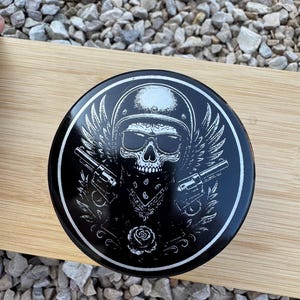 Könnte beinhalten: Ein schwarzer runder Behälter mit einer weißen Grafik eines Schädels mit Helm und Sonnenbrille, mit Flügeln, zwei Pistolen und einem Bandana. Am unteren Rand des Designs befindet sich eine Rose.