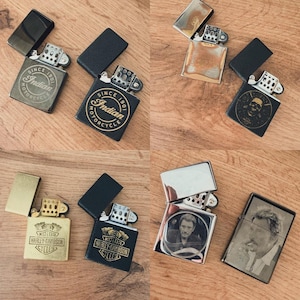 May include: A selection of metal lighters, some open, some closed, with diverse designs. The lighters display logos such as "Indian Motorcycle" and "Harley-Davidson", a skull, and portraits of a man. The lighters are on a wooden surface.