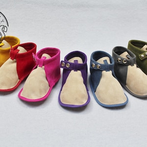 Könnte beinhalten: Eine Auswahl an Baby-Booties in verschiedenen Farben: Gelb, Rot, Pink, Lila, Blau, Grau und Olivgrün. Jeder Bootie hat ein beigefarbenes, Wildleder-ähnliches Fußbett und ein farblich abgesetztes Oberteil mit Riemen.