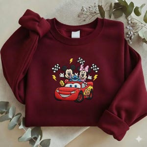 May include: Burgundy sweatshirt featuring an embroidered design of Mickey Mouse, Minnie Mouse, Stitch, and Lightning McQueen. The design includes checkered flags and lightning bolts. The sweatshirt has a crew neck and long sleeves.