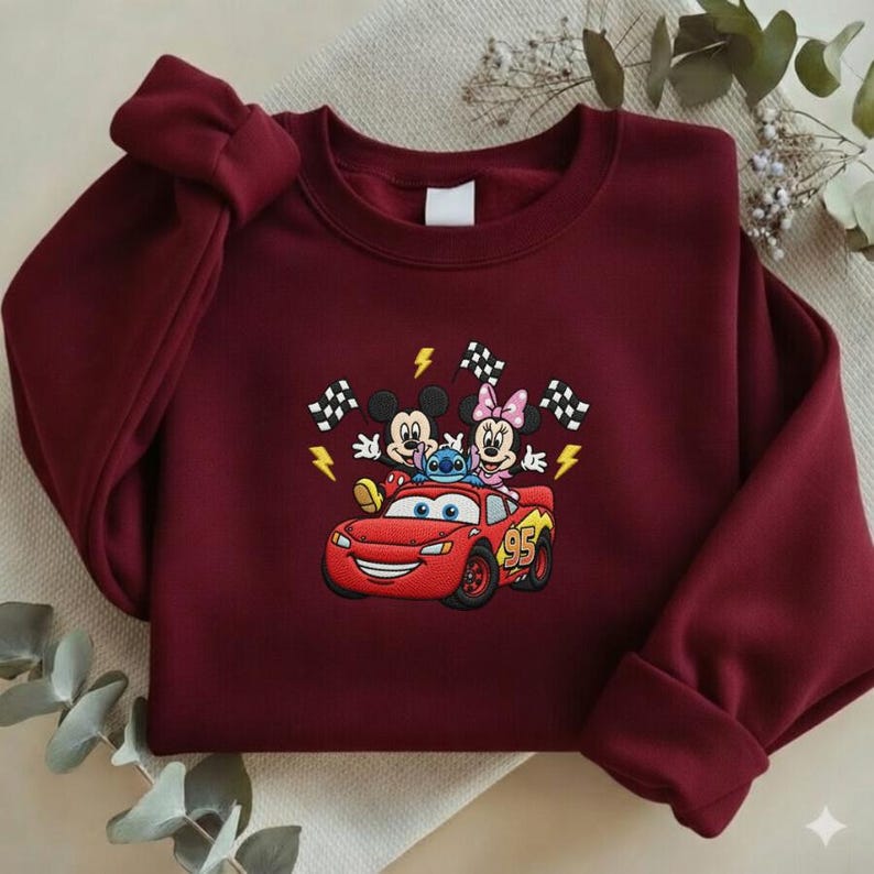 May include: Burgundy sweatshirt featuring an embroidered design of Mickey Mouse, Minnie Mouse, Stitch, and Lightning McQueen. The design includes checkered flags and lightning bolts. The sweatshirt has a crew neck and long sleeves.