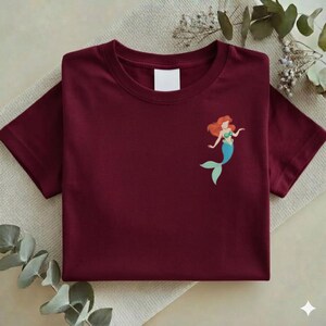 May include: A maroon t-shirt featuring a small graphic of a mermaid with red hair, a green top, and a blue tail. The shirt is folded neatly on a neutral-colored surface, with a blank white tag near the neckline.