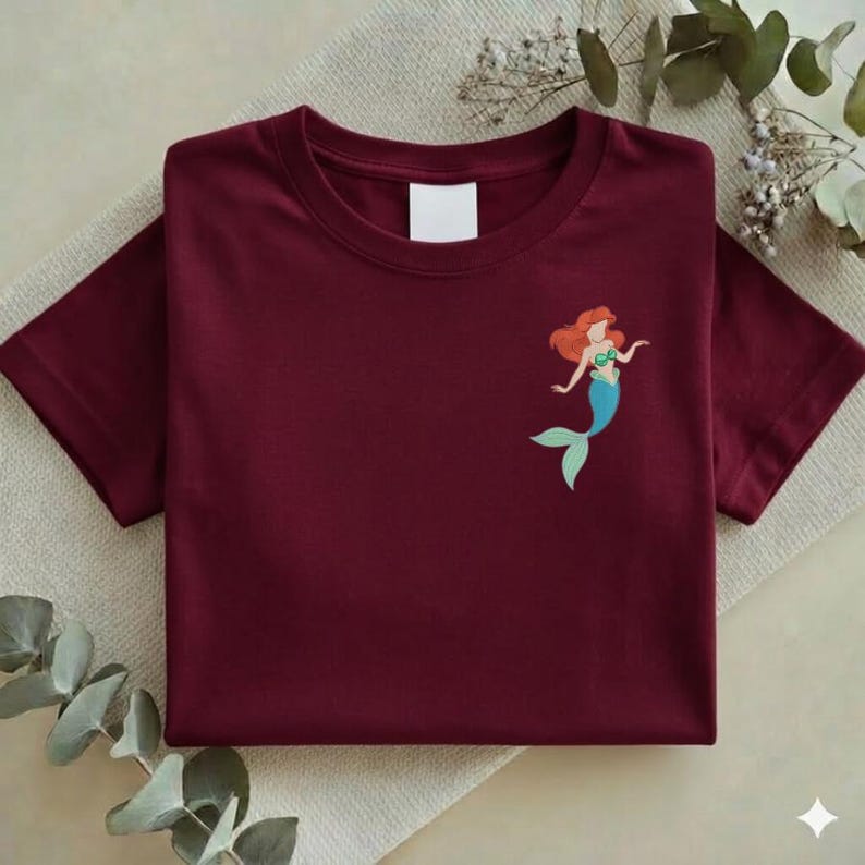 May include: A maroon t-shirt featuring a small graphic of a mermaid with red hair, a green top, and a blue tail. The shirt is folded neatly on a neutral-colored surface, with a blank white tag near the neckline.