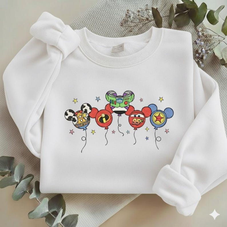 Może przedstawiać: Biała bluza z wyszywanymi balonami z postaciami z kresk&oacute;wek. Balony przedstawiają Myszka Miki, Buzz Lightyear, logo Iniemamocnych, Zygzaka McQueena i gwiazdę. Każdy balon jest w innym kolorze i ma pod spodem gwiazdkę.