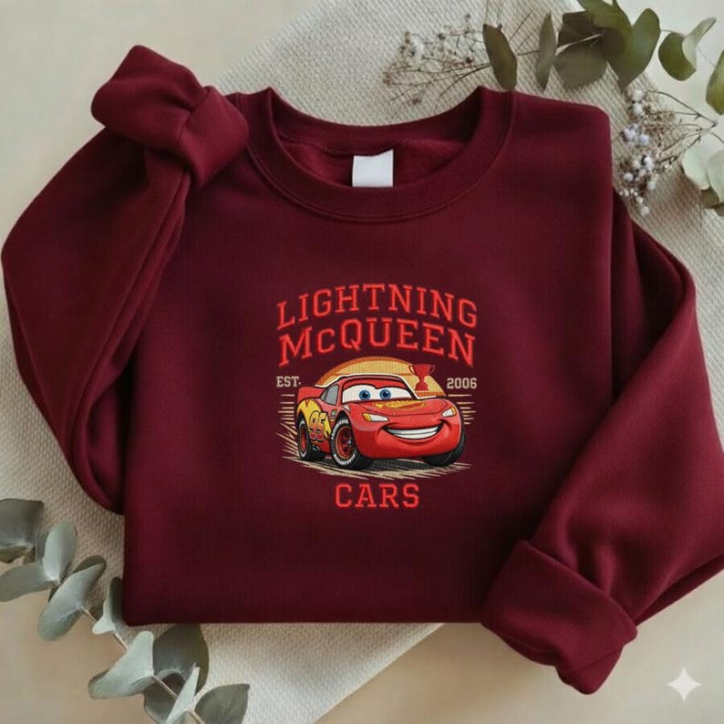 Puede incluir: Una sudadera burdeos con un dise&ntilde;o de Rayo McQueen de la pel&iacute;cula Cars. El dise&ntilde;o incluye el texto "Lightning McQueen" encima de la imagen del coche, con "Est. 2006" y "Cars" debajo. La sudadera tiene cuello redondo y mangas largas.
