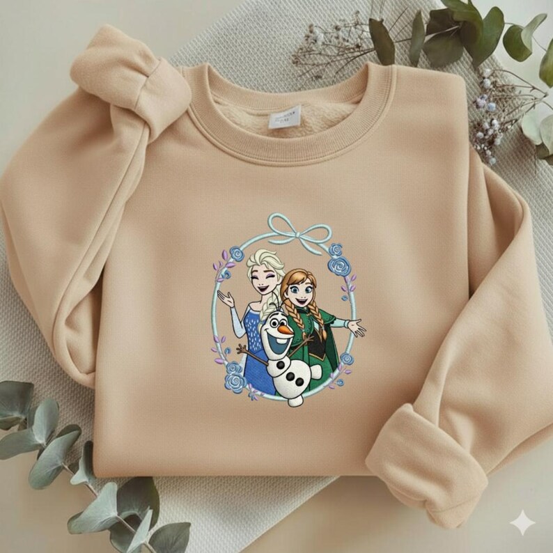 Puede incluir: Sudadera beige con un gr&aacute;fico colorido de Elsa, Anna y Olaf de la pel&iacute;cula Frozen, rodeados por una corona floral. La sudadera tiene cuello redondo y mangas largas con pu&ntilde;os acanalados.