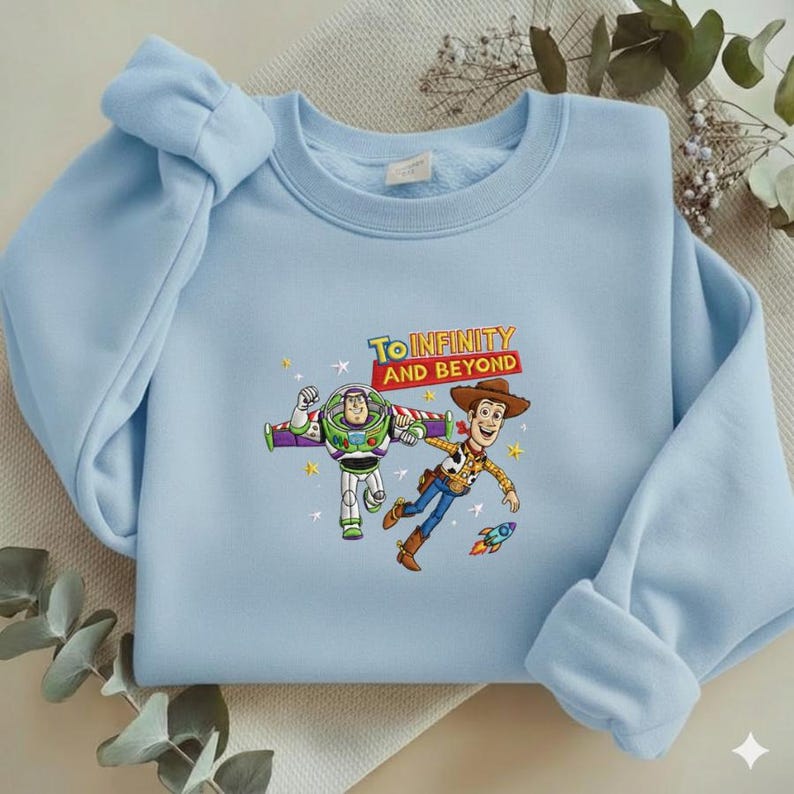 K&ouml;nnte beinhalten: Hellblaues Sweatshirt mit Buzz Lightyear und Woody aus Toy Story, mit dem Text "To Infinity and Beyond". Die Grafik zeigt die Charaktere mit einer Rakete und Sternen. Das Sweatshirt hat einen Rundhalsausschnitt und lange &Auml;rmel.